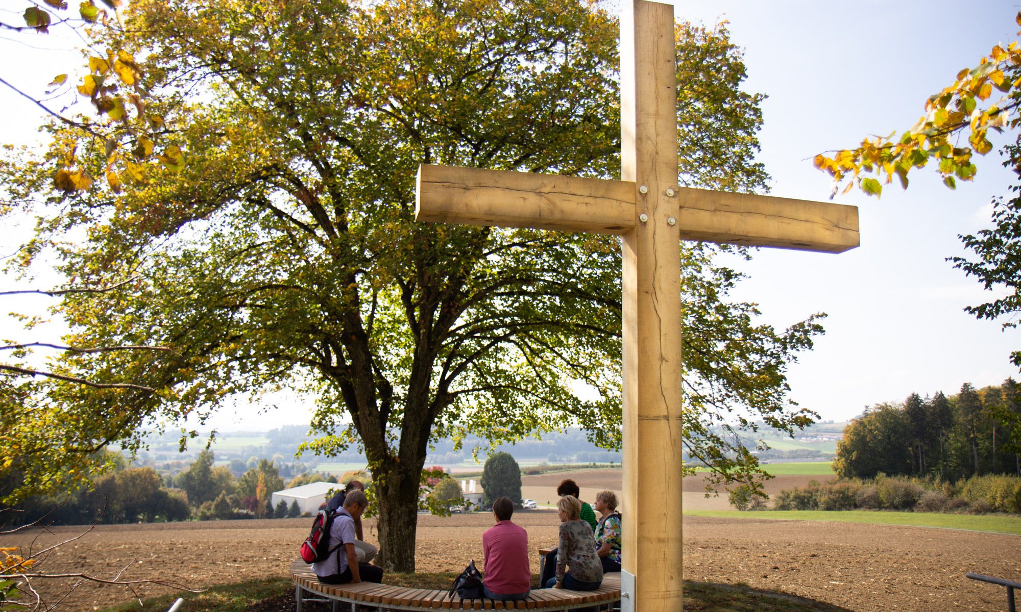 Station 5 des Vaterunser-Weges mit dem Kreuz im Rücken. Davor Menschen auf der Bank.