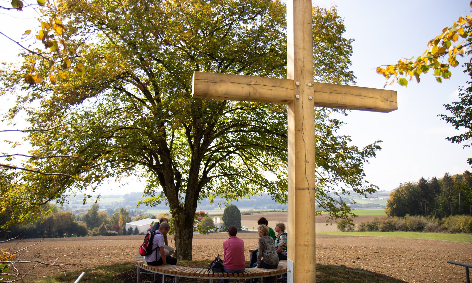 Station 5 des Vaterunser-Weges mit dem Kreuz im Rücken. Davor Menschen auf der Bank.